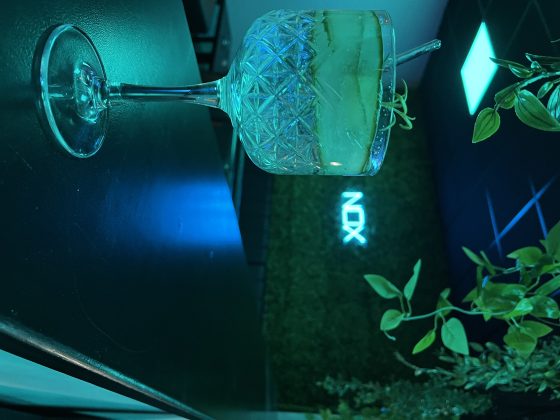 Cocktailglas an beleuchteter Bar mit Pflanzen und NOX-Schild.